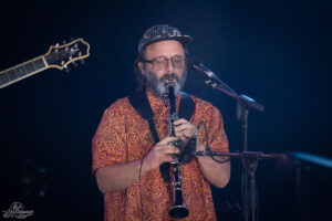 Musician wearing a patterned orange shirt and a patterned cap playing a clarinet on a dimly lit stage, with a microphone nearby and a guitar neck to the left.