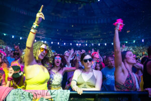 Crowd at a concert raising arms and dancing, colorful lights illuminating the stadium.