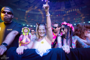 Woman in white top raises her hand in the crowd at a neon-lit concert, smiling with friends around her.