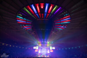 Concert at Madison Square Garden featuring a circular rainbow-lit ceiling and a bright multi-screen stage with a cheering crowd.
