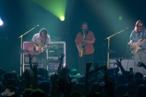Live band on stage: guitarist left, saxophonist center, bassist right; green/blue lights and cheering crowd.
