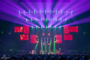Concert stage with purple and teal lights, LED panels above and musicians performing to a large crowd.