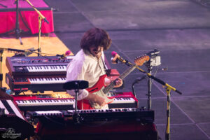 Musician in a white shirt smiling while playing a red electric guitar among stacked keyboards on a dimly lit stage with microphones nearby