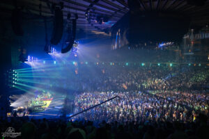 Large indoor concert venue with a band performing on stage left, blue and green spotlights sweeping over a packed audience.
