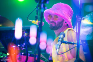 Drummer with pink fluffy hat and yellow vest performing on a neon-lit stage with a microphone nearby.