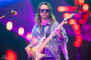Musician wearing sunglasses and a denim jacket plays a light-colored bass on a neon-lit stage.