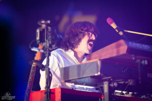 Musician with glasses and mustache singing loudly at a keyboard on stage, purple lighting in the background.