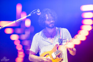 Male guitarist playing a sunburst electric guitar on stage under blue and pink lights, wearing a patterned shirt and necklace.