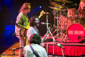 Rock band performing on stage; lead singer with guitar at a mic, drummer behind a red 'BIG DRUM!' bass drum.