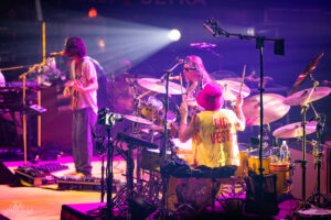 Drummer in a bright yellow vest with 'BIG VEST' and a pink hat plays behind a drum kit on a neon-lit stage.