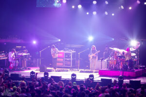 Live band on stage performing with keyboards, guitars, bass, and drums under purple stage lights, crowded audience in front.