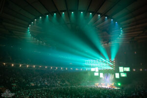 Large indoor concert with teal stage lights and a full audience facing a brightly lit stage.