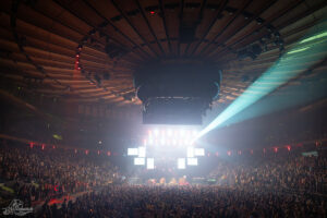 Massive indoor concert at Madison Square Garden with a packed arena and a bright, rectangular stage setup amid dramatic overhead lighting.