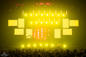 Concert stage bathed in bright yellow lights with rectangular screens and a cheering crowd in front of the stage.