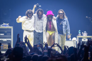 Four male performers pose together on a concert stage as the crowd cheers in front of them.