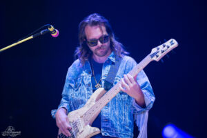 Bassist with long hair and sunglasses wearing a denim jacket, playing a cream-colored bass on stage.