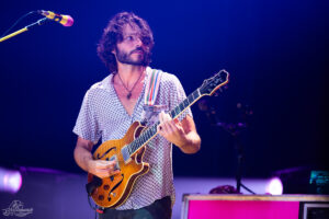 Male guitarist with curly hair in a patterned shirt playing a sunburst hollow-body electric guitar on a dimly lit stage during a live concert, with a microphone nearby.