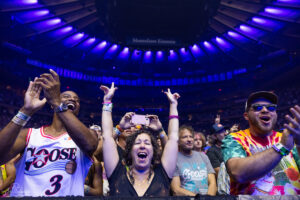 Crowd at a concert cheering with hands in the air; left man in a Goose basketball jersey, center woman with glitter, right man in a tie-dye shirt and sunglasses under purple lights.