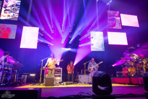 Band performs on a brightly lit stage with purple and blue lights, guitarist center, keyboardist left, bassist right, and a drummer on the far right, with large video screens overhead.