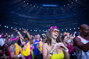 Smiling woman in a yellow top with a pink flower crown and orange sunglasses at a crowded concert in a large arena.