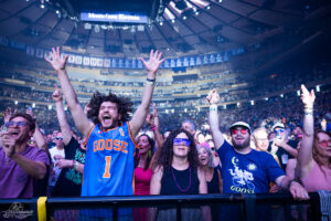 Crowd at a concert in a large arena, man in a blue Goose jersey with number 1 raises his arms in excitement as others cheer around him.