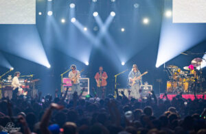 Band performing on a lit stage with guitar, saxophone, bass, and drums, crowd raising hands in front of them under bright blue spotlights.