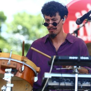 Male musician in a purple shirt wearing round sunglasses, focused on playing drums at an outdoor performance with a keyboard nearby.