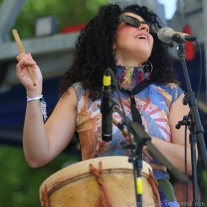 Female singer performing outdoors, wearing sunglasses and a colorful top, singing into a microphone while playing a hand drum.
