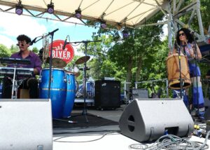 Musicians performing on an outdoor stage with drums, keyboard, and conga; left musician in purple shirt plays keyboard, right musician wears sunglasses and sings into a microphone wearing colorful clothing and stands by conga drums.