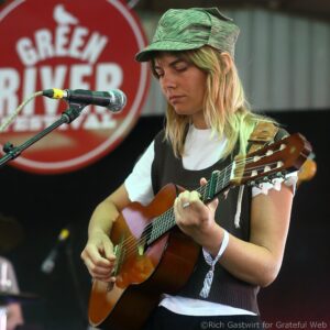 Woman musician wearing a green cap, playing an acoustic guitar and singing into a microphone on stage with a red backdrop behind her.