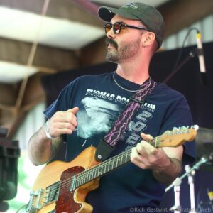 Guitarist on stage wearing a green cap and sunglasses, playing a natural wood electric guitar with a patterned strap.