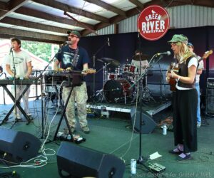 Band performing on a stage at the Green River Festival; guitarist and singer front center, drummer in back, sign above reads Green River Festival.