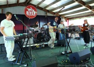 Band performing on an outdoor stage at the Green River Festival with guitars, drums, and keyboards visible.