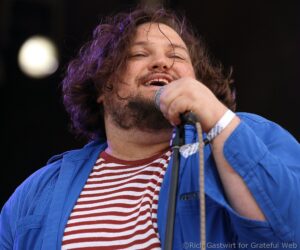 Male singer performing on stage, close-up with microphone, wearing a blue jacket and striped shirt, facial hair and purple lighting in the background