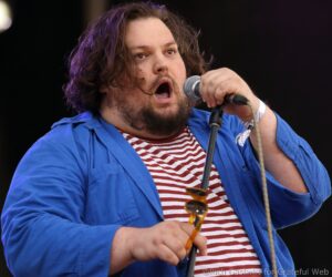 Male singer mid-performance holding a microphone, wearing a blue shirt over a red-and-white striped tee, with sunglasses hanging from his chest.