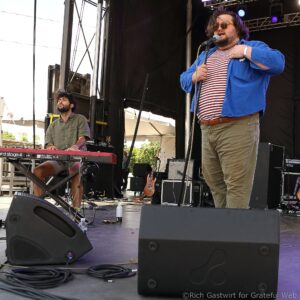 Two male musicians perform on an outdoor stage: a keyboardist on the left and a bearded singer in a blue jacket and striped shirt singing into a microphone on the right.