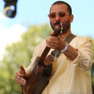 Musician outdoors wearing sunglasses and a beige shirt, holding and playing a guitar with a strap visible across the chest.