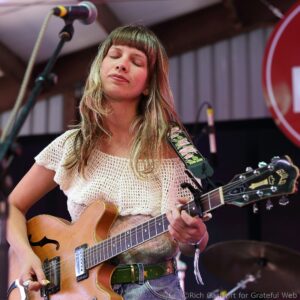 Female musician with long hair and closed eyes playing a sunburst electric guitar on stage, wearing a cream crocheted top and a floral strap.]","Guitarist performing on stage, close-up of hands on the neck of a sunburst guitar with a floral strap.