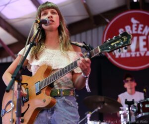 Female musician with fringe sings into a microphone while playing an electric guitar on stage, wearing a cream knit top and a decorative belt, with a red circular sign in the background.