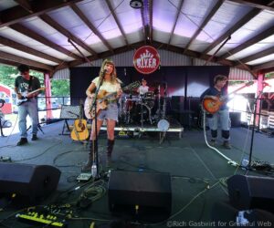 Female singer with an acoustic guitar performing on a festival stage, with a drummer and two guitarists behind her and a Green River Festival sign above.