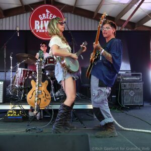 Two musicians perform on an outdoor stage: a woman with a guitar facing left and a tattooed man with a bass facing her, drums and amps in the background, and a red round sign behind them