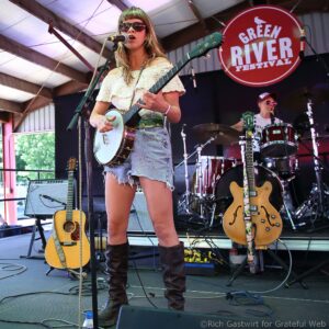 Female musician sings into a microphone while playing a light-colored resonator guitar on stage, with a 'Green River Festival' sign in the background.
