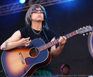 Female guitarist performing on stage, wearing sunglasses and a chunky chain necklace, playing a sunburst acoustic guitar.