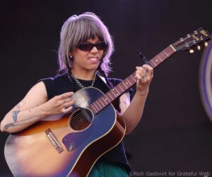 Guitarist with short gray hair and sunglasses playing a sunburst acoustic guitar on stage, during a live performance.