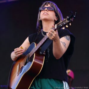 Person playing an acoustic guitar on stage, wearing dark sunglasses and a silver chain.