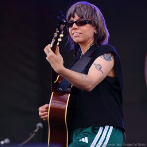 Female guitarist performing on stage, wearing sunglasses and a black outfit, strumming a brown acoustic guitar.
