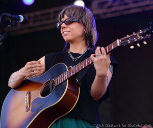 Female guitarist performing on stage with an acoustic guitar, wearing sunglasses and a silver chain necklace, tattoos visible on arms