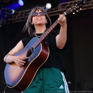 Female guitarist on stage wearing sunglasses, playing a sunburst acoustic guitar and singing into a microphone.