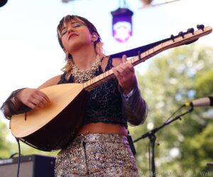 Female guitarist performing on stage, eyes closed, playing an acoustic guitar outdoors.