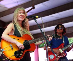 Female musician singing into microphone while playing a sunburst acoustic guitar on stage, with a second guitarist in a red guitar nearby.